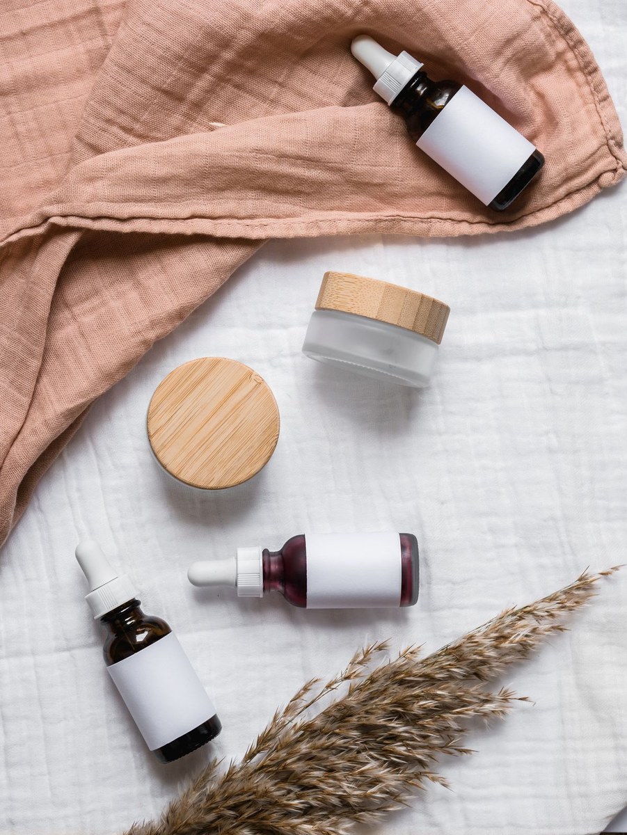 Unlabeled serum bottles and cream jars flat-laid on fabric with dried grass