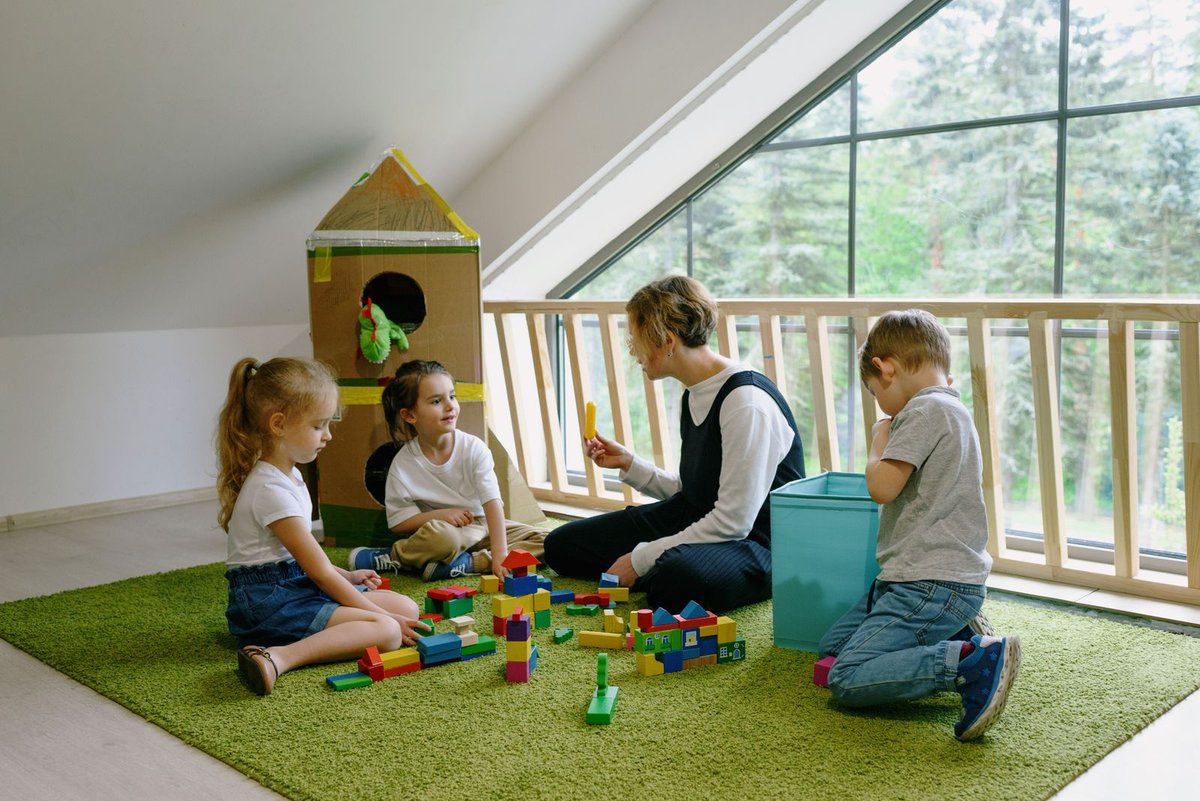 Teacher and three children building with colourful wooden blocks on a green carpet in a bright playroom