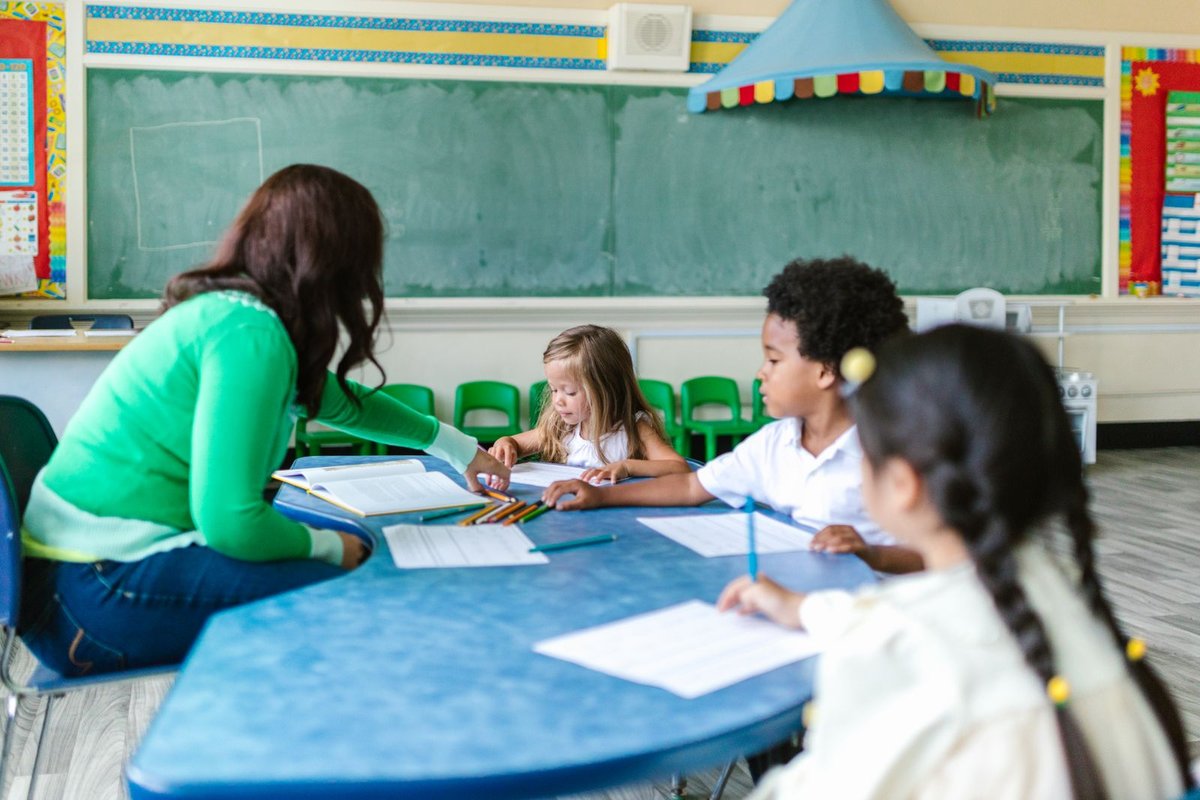 Teacher in a green shirt guiding three young children working with books and pencils at a blue classroom table